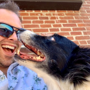 To the left is a white man with spiky light brown hair and closely shaved mustache and beard wearing reflective sunglasses and alight blue button shirt with small green tropical print dispersed pattern is outside by a red brick wall. His mouth is open in a wide smile. The man is looking at a medium-length hair dog located to the right foreground. The dog has black fur on the head and ears and a spot of white on end of its nose, plus a white chin and neck. The dog is looking at the man and has its mouth open showing clean white teeth.