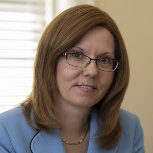 Josipa-Roksa-sq-crop A slightly smiling white woman showing a bit of her upper front teeth with light red hair, neatly trimmed eyebrows, and brown eyes wearing copper framed glasses with rounded oblong lenses. She is wearing a silver necklace with rectangular chain links and a light blue blazer with black piping trim around the collar/lapel. She is inside in front of a window with horizontal blind and beige wall.