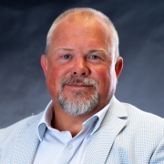 A smiling white man with blue eyes and a sun-kissed complexion, crew cut blond hair, shortly trimmed salt and pepper colored mustache and goatee wearing a white button shirt with collar and small, blue checked pattern and a white, gold, and blue checked blazer posed in front of a shades of blue and grey portrait backgound.