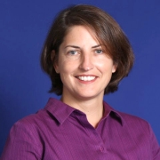 A smiling white woman with light eyes and short brown bob haircut that is parted on the left an angled across the edge of her right eye and brow. She is wearing a purple collared button shirt and is facing forward looking directly into the camera in front of a dark blue background.