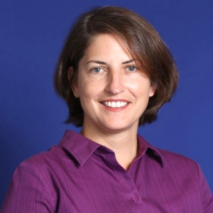 yvette-pearson-sq A smiling white woman with light eyes and short brown bob haircut that is parted on the left an angled across the edge of her right eye and brow. She is wearing a purple collared button shirt and is facing forward looking directly into the camera in front of a dark blue background.