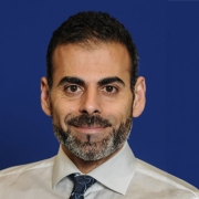 A smiling man with medium toned skin and dark eyes has dark short cut hair, mustache and salt and pepper beard wearing a collared ivory shirt with dark patterned tie.